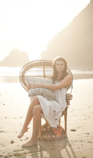 Carley Kahn "Checker" pillow case. Model is sitting in peacock chair on beach and holding onto pillow. 